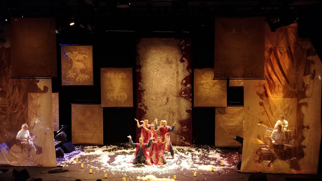 Performers pose on a stage surrounded by large paper panels and scattered white material