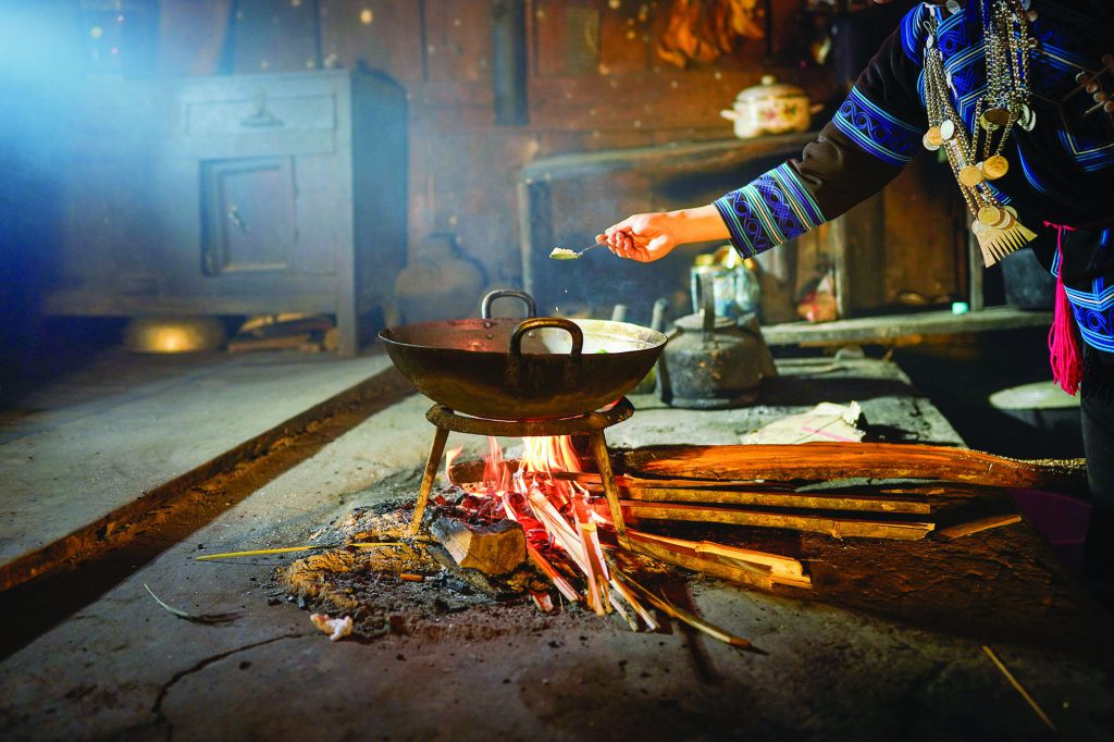 Ha Nhi woman cooking over an open fire inside a traditional kitchen, stirring food in a wok