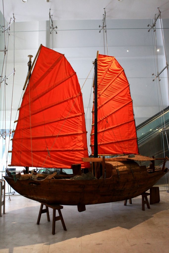 A traditional wooden boat with vibrant red sails is displayed indoors on stands