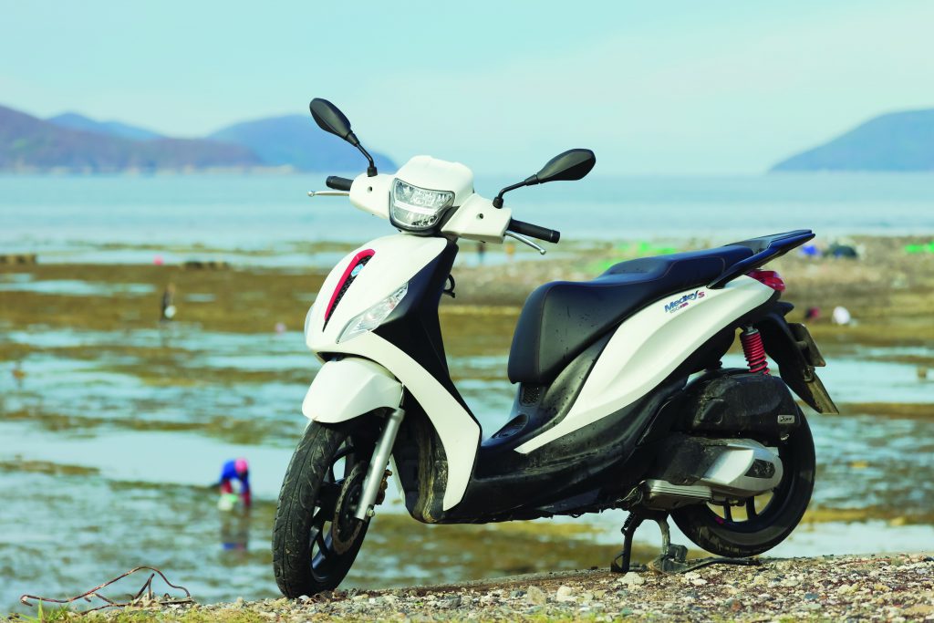 Xe tay ga Piaggio Medley S đỗ bên bờ biển, phía xa là mặt nước và dãy núi