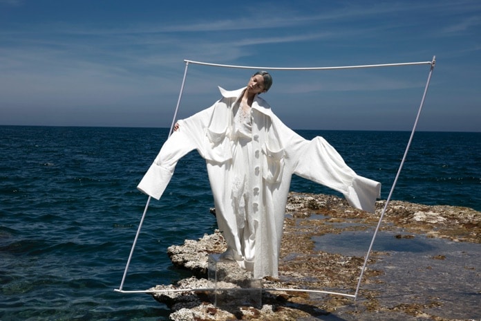 Model in an oversized white coat with dramatic sleeves posing on a rocky outcrop by the deep blue sea.