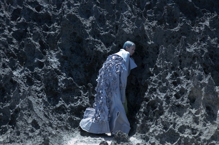 Fashion model in a textured grey coat walking through a rugged landscape of dark volcanic rocks.