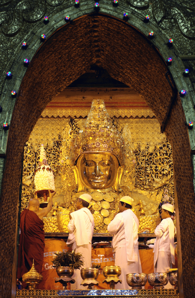 A golden Buddha face is framed by an ornate archway with monks attending it