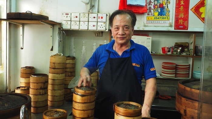 Người đàn ông lớn tuổi mỉm cười đứng trước các giỏ hấp bánh bao tại một cửa hàng