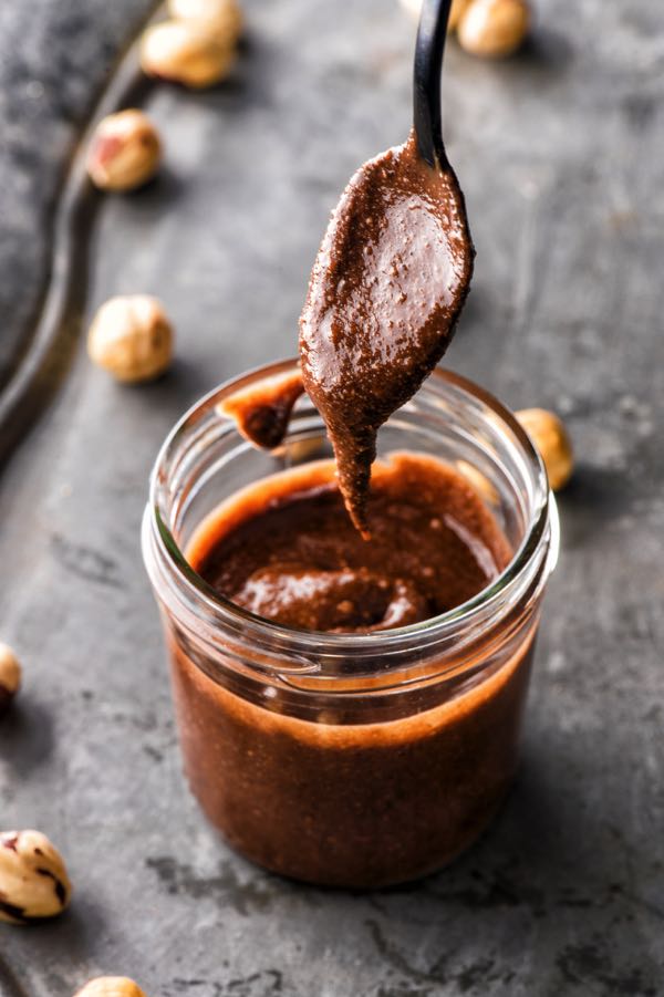 Lọ bơ hạt dẻ socola sánh mịn với chiếc thìa đang múc lên đầy hấp dẫn trên nền xám.