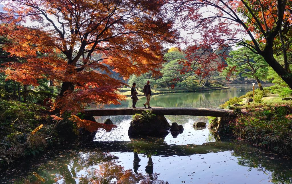 Two people walk across a small wooden bridge over calm water surrounded by autumn trees