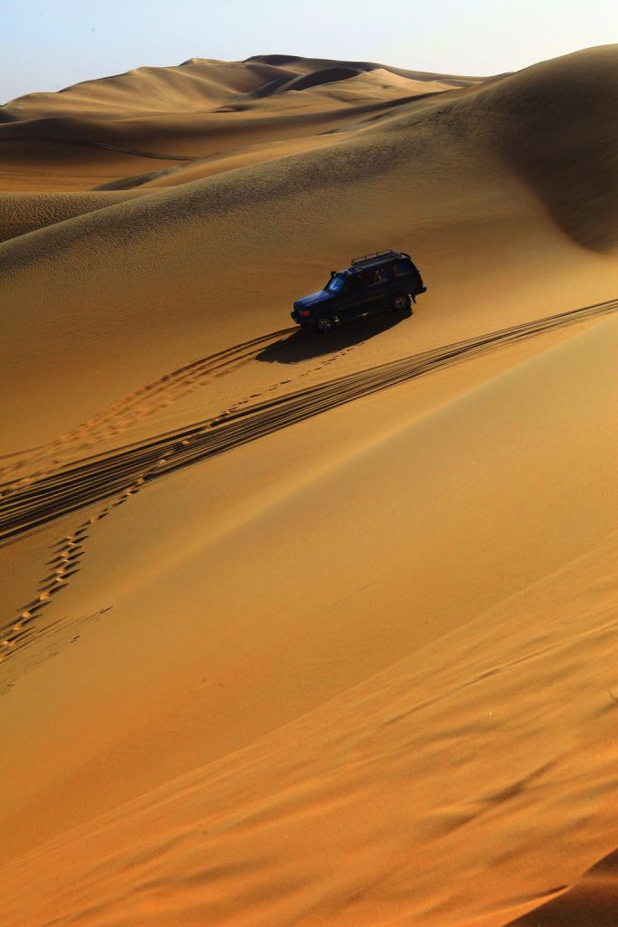 A dark vehicle drives across rolling golden sand dunes under a clear sky