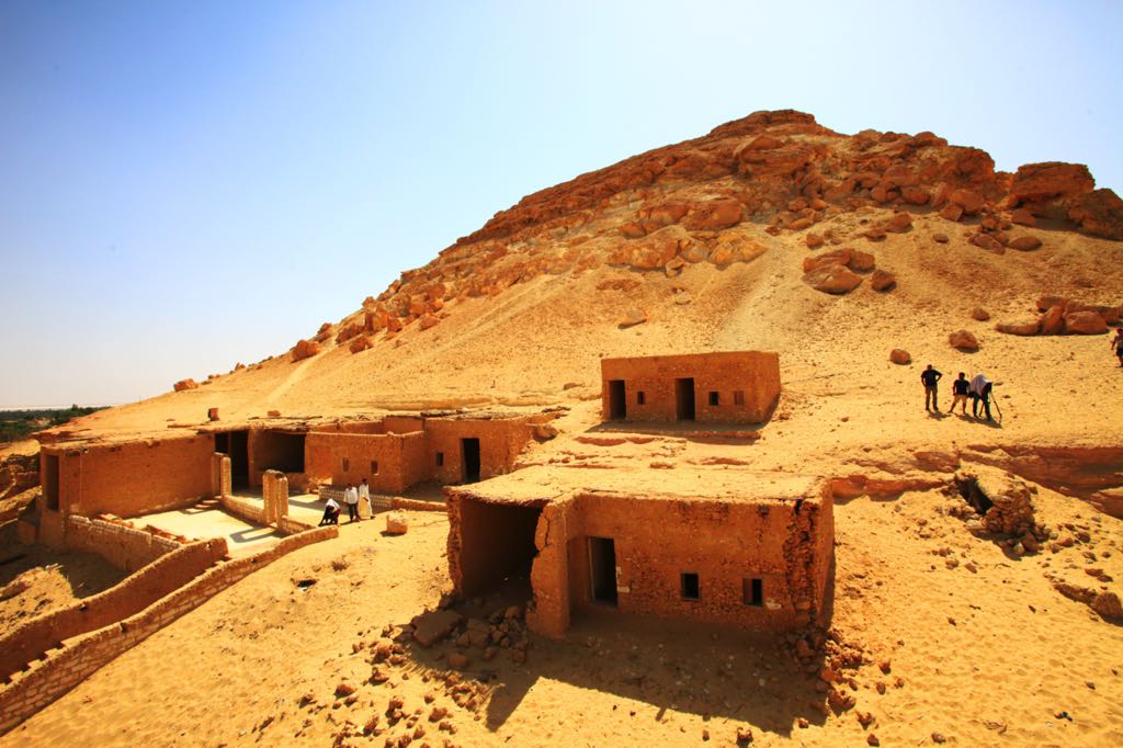 Mud brick buildings sit below a large sandy hill under a bright blue sky