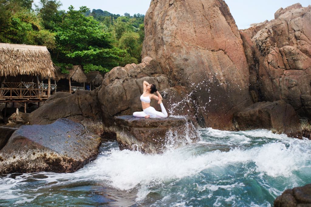 A woman performing a pigeon yoga pose on a rock as ocean waves splash behind her