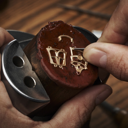 Bàn tay đang lắp ráp chi tiết vàng hình chữ samurai lên mặt đồng hồ màu nâu sẫm