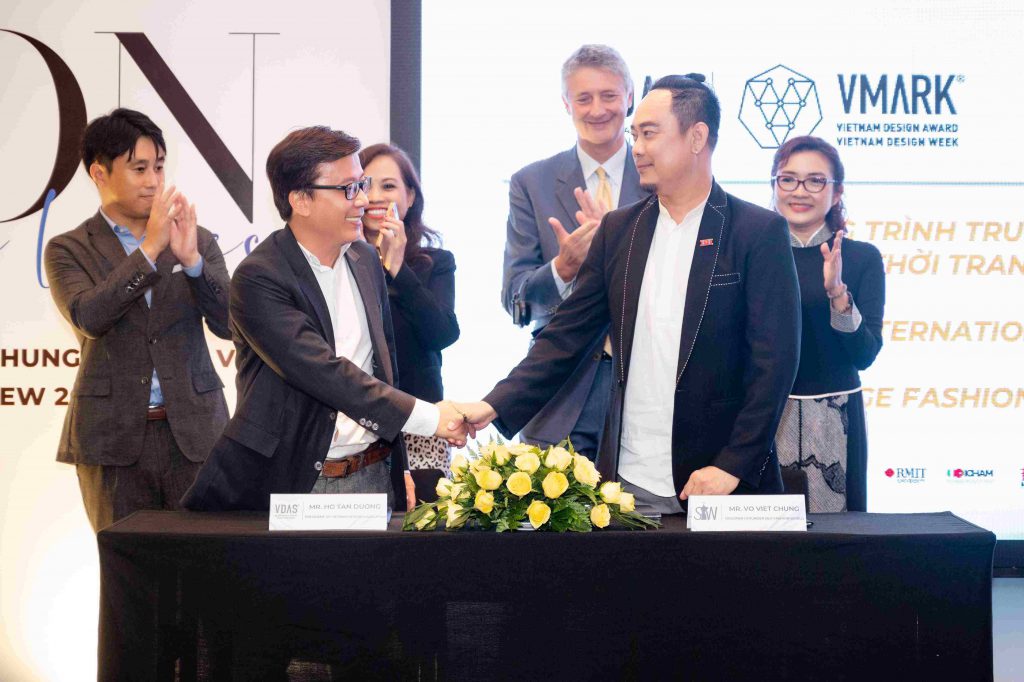 Hai người đàn ông bắt tay trên sân khấu trước khán giả và backdrop sự kiện thiết kế thời trang