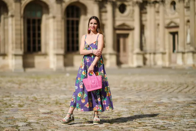Cô gái mặc váy hoa cầm túi xách hồng đứng trước tòa nhà kiến trúc cổ kính