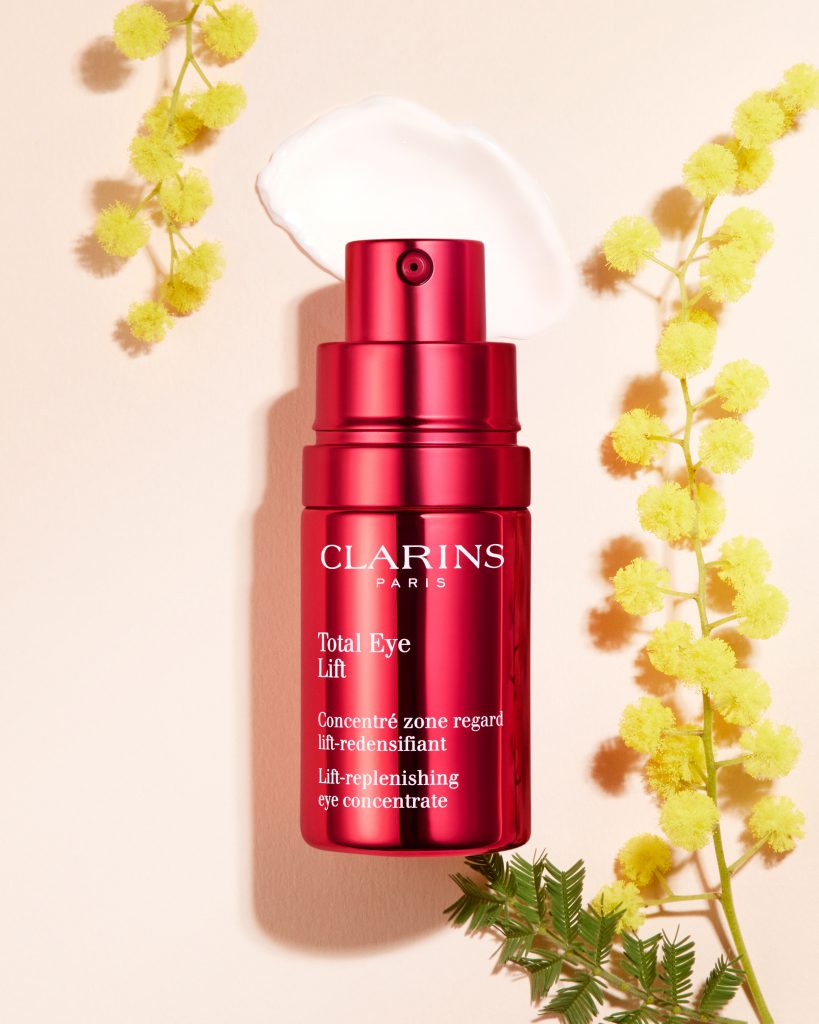 Sản phẩm kem dưỡng mắt Clarins Total Eye Lift màu đỏ nằm giữa các nhánh hoa mimosa vàng trên nền hồng nhạt