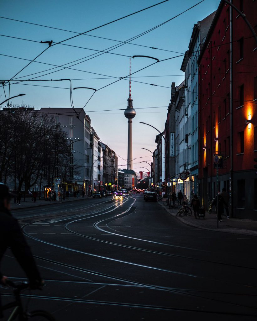 A Berlin street scene features the TV tower and tram tracks under a blue sky