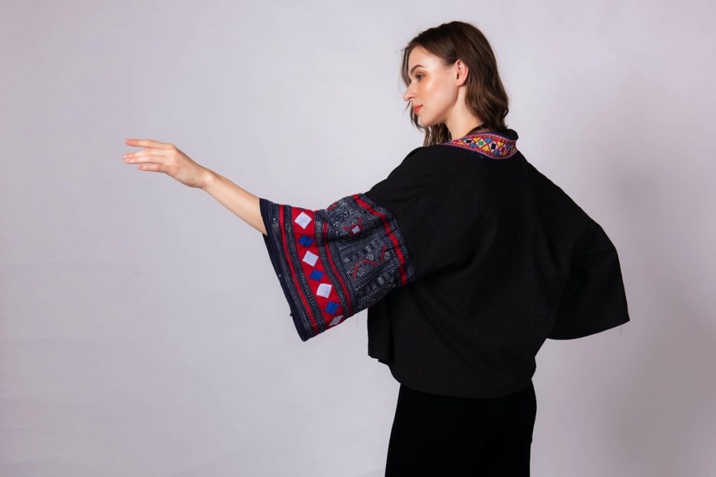 Người phụ nữ mặc áo khoác đen tay loe họa tiết thổ cẩm nổi bật trên nền trắng