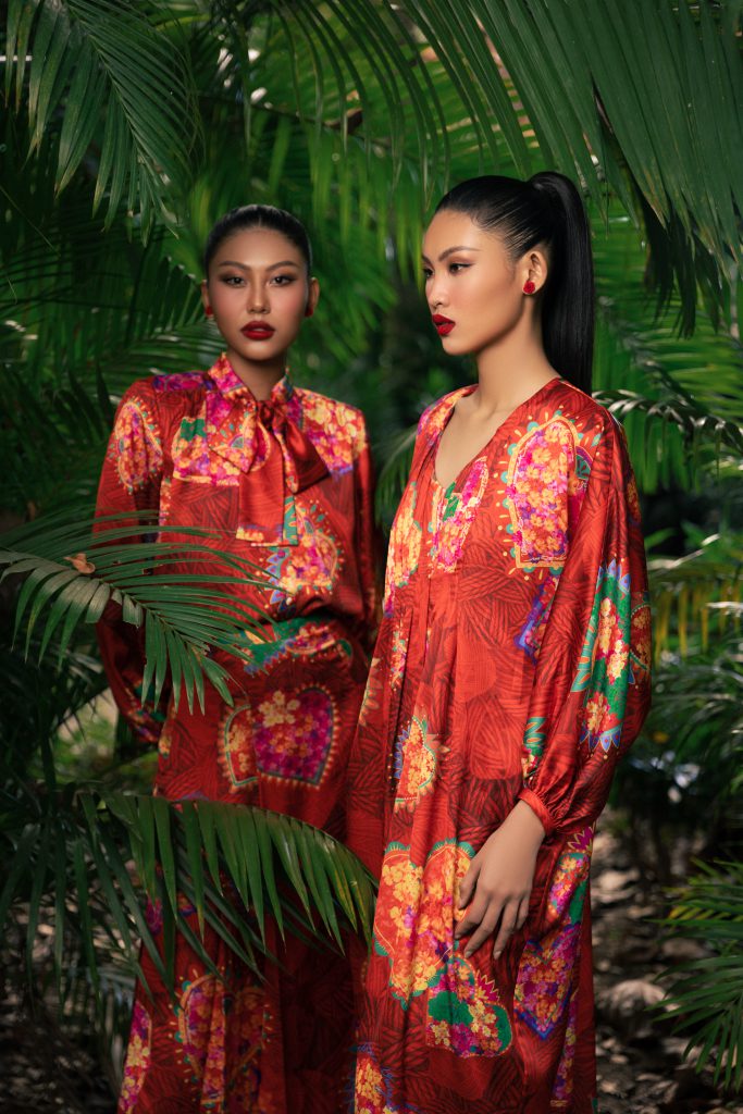 Hai người mẫu diện trang phục màu đỏ hoa văn nổi bật giữa khung cảnh lá xanh