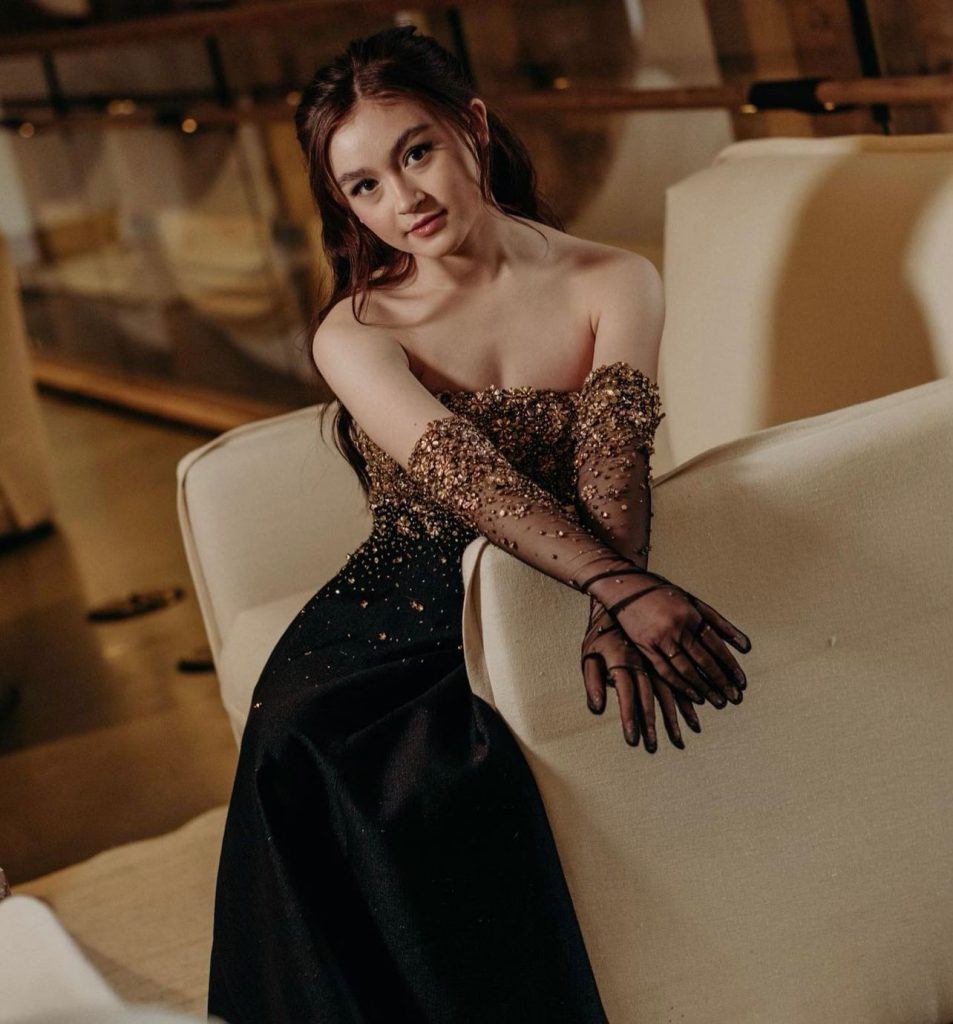 Người mẫu diện váy đen lộng lẫy cùng găng tay xuyên thấu tinh tế đang ngồi trên ghế sofa
