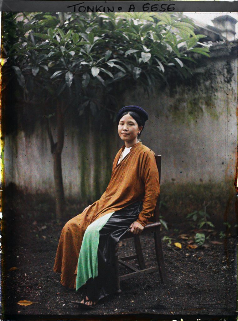 Người phụ nữ mặc áo dài và quần đen ngồi trên ghế gỗ trước cây xanh