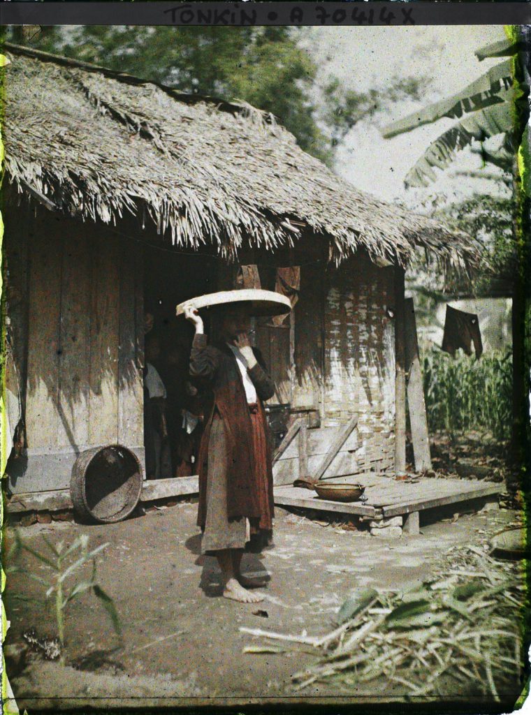 Người phụ nữ đội thúng bên ngoài một ngôi nhà mái tranh ở Tonkin