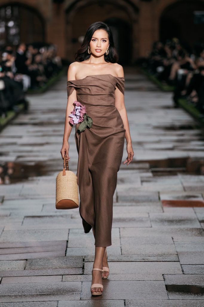 Người mẫu trình diễn váy trễ vai màu nâu cùng giỏ xách mây trên sàn diễn thời trang