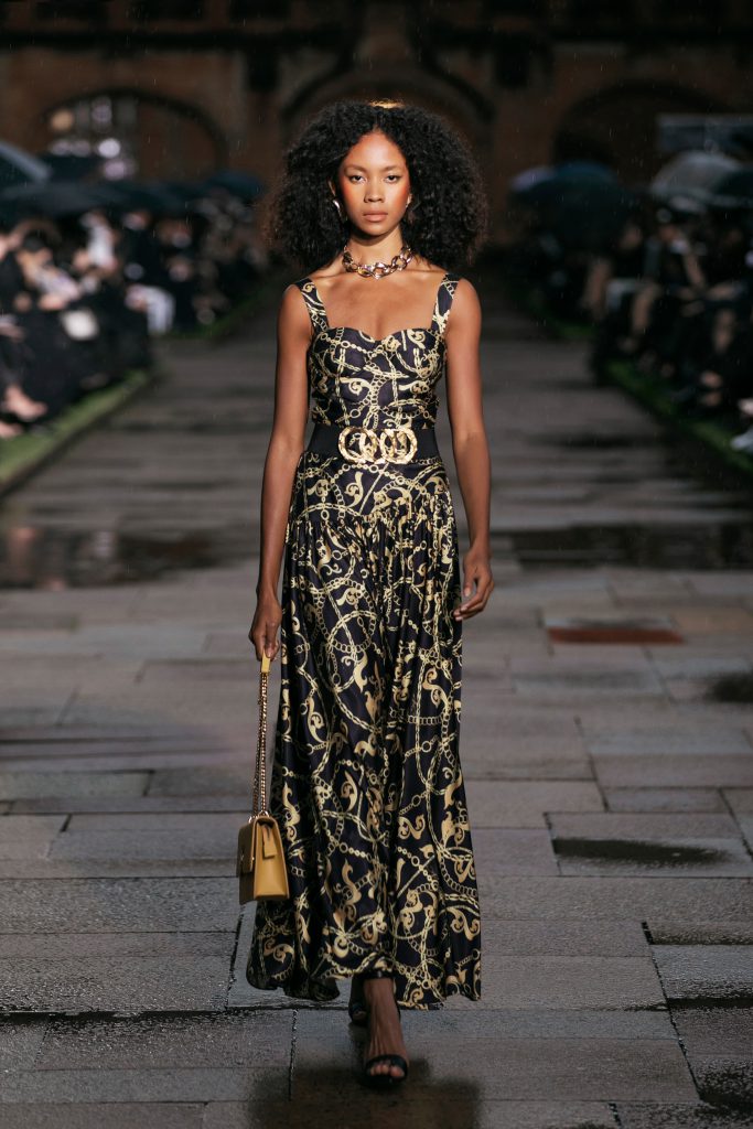 Người mẫu trình diễn váy dài họa tiết xích vàng trên sàn diễn thời trang ngoài trời