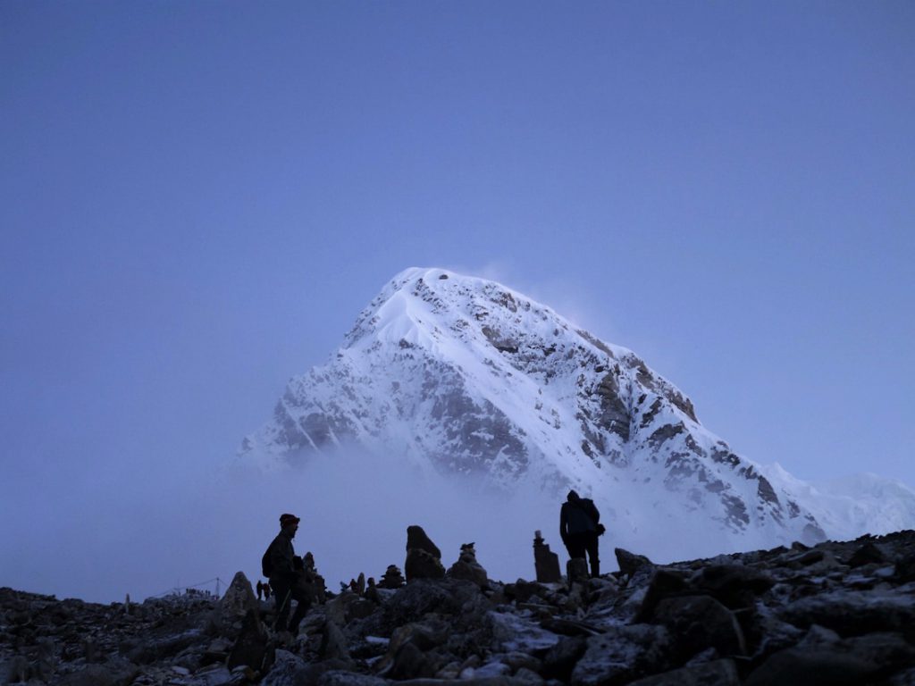 Silhouetted figures stand before a large snow covered mountain under a blue sky