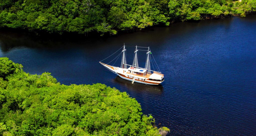 A white and brown sailing ship travels down a dark river surrounded by lush green trees