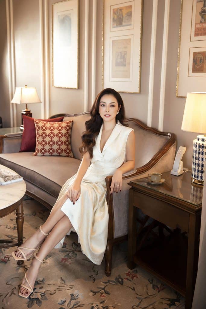 Jennifer Phạm diện váy trắng tinh tế ngồi trên ghế gỗ chạm khắc