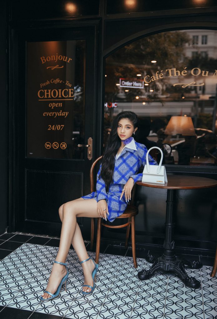 Á hậu Minh Kiên ngồi duyên dáng trước cửa quán cà phê với trang phục màu xanh nổi bật