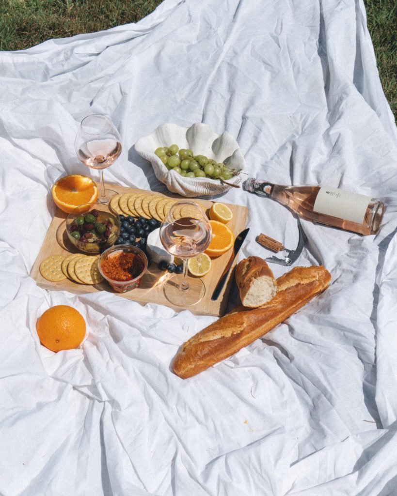 A picnic blanket displays wine glasses fruit cheese and a baguette