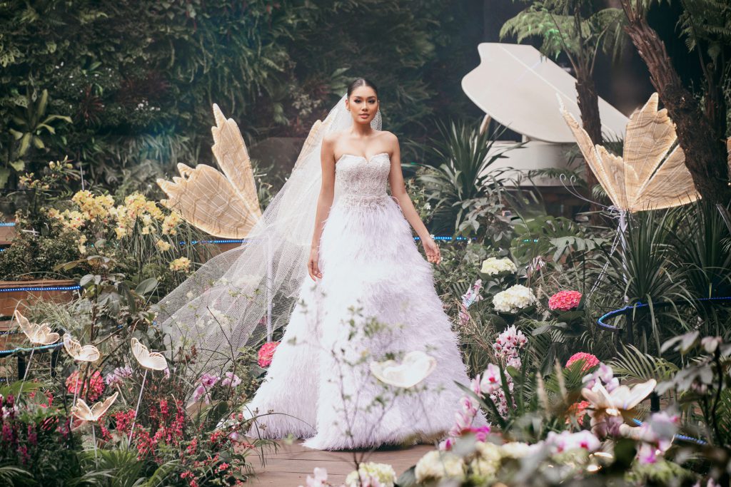 Người mẫu diện váy cưới trắng tinh khôi sải bước giữa không gian trang trí hoa lá