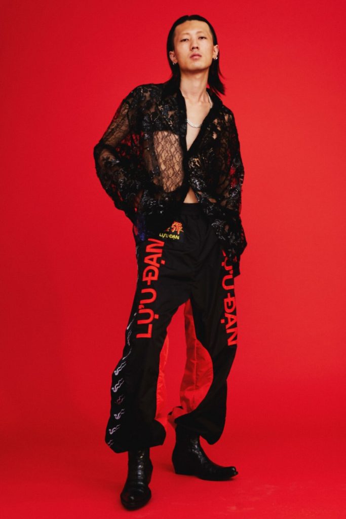 Người đàn ông mặc trang phục đen họa tiết hoa và quần rộng in chữ trên nền đỏ