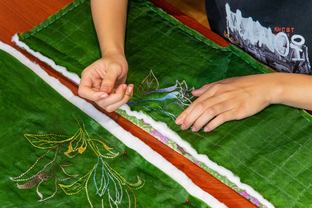 Bàn tay đang thêu hoa lá tỉ mỉ trên nền vải chần bông màu xanh lá cây