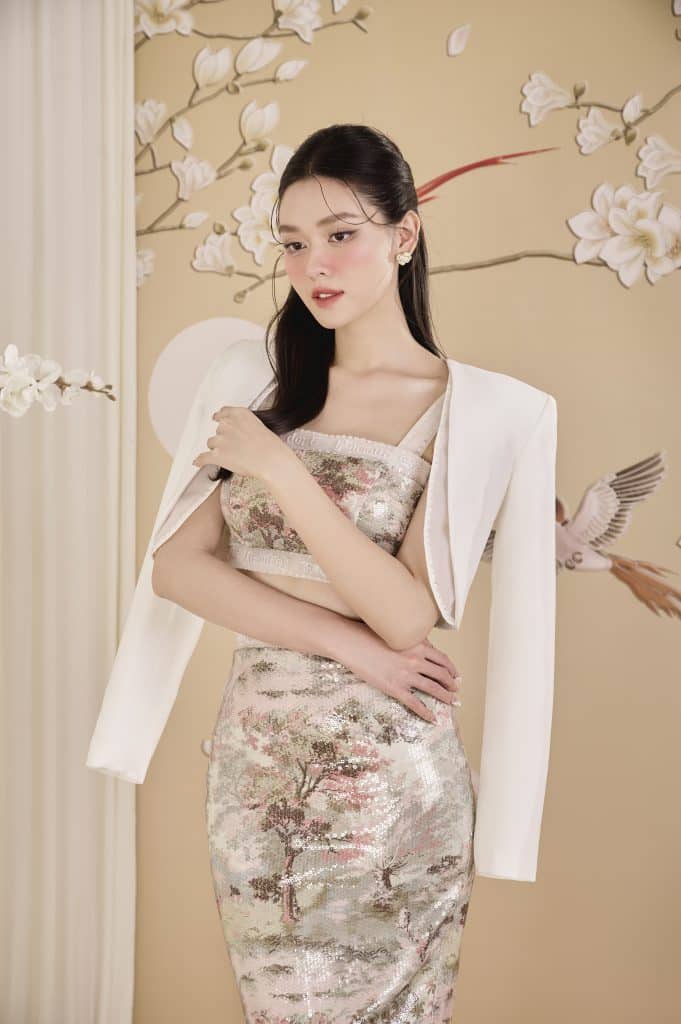 Người mẫu mặc váy lấp lánh và áo khoác trắng tạo dáng trước nền hoa mai