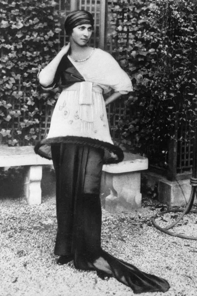 A woman in a long gown and headwrap poses beside a stone bench in a garden