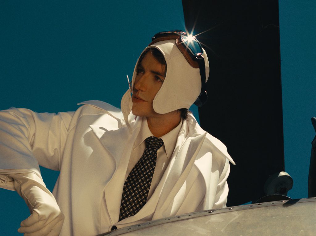 Người mẫu nam mặc áo khoác trắng đội mũ phi công và đeo kính râm