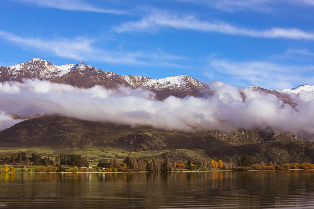 Snowy mountains rise above low clouds reflecting in a still lake with autumn trees