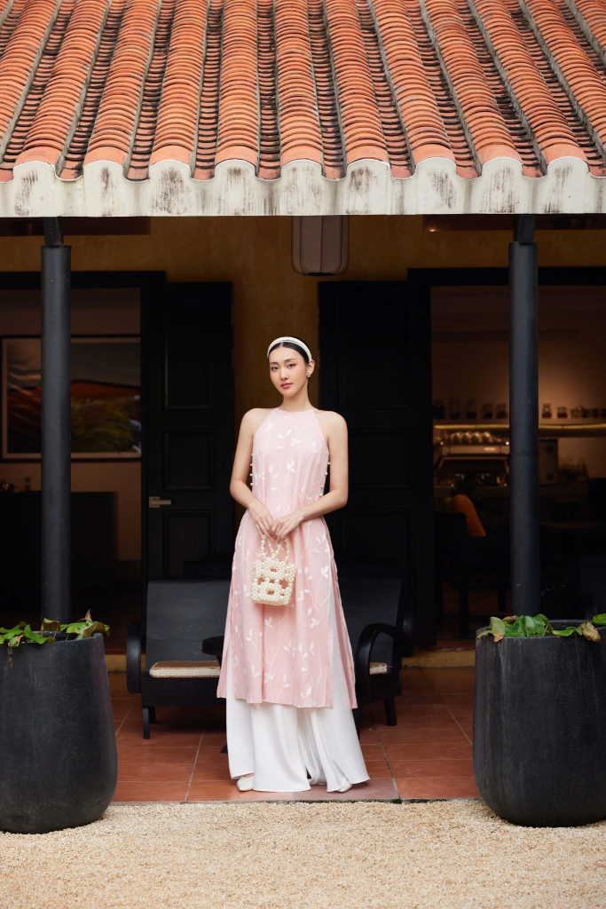 Người phụ nữ mặc áo dài hồng đứng trước một ngôi nhà mái ngói