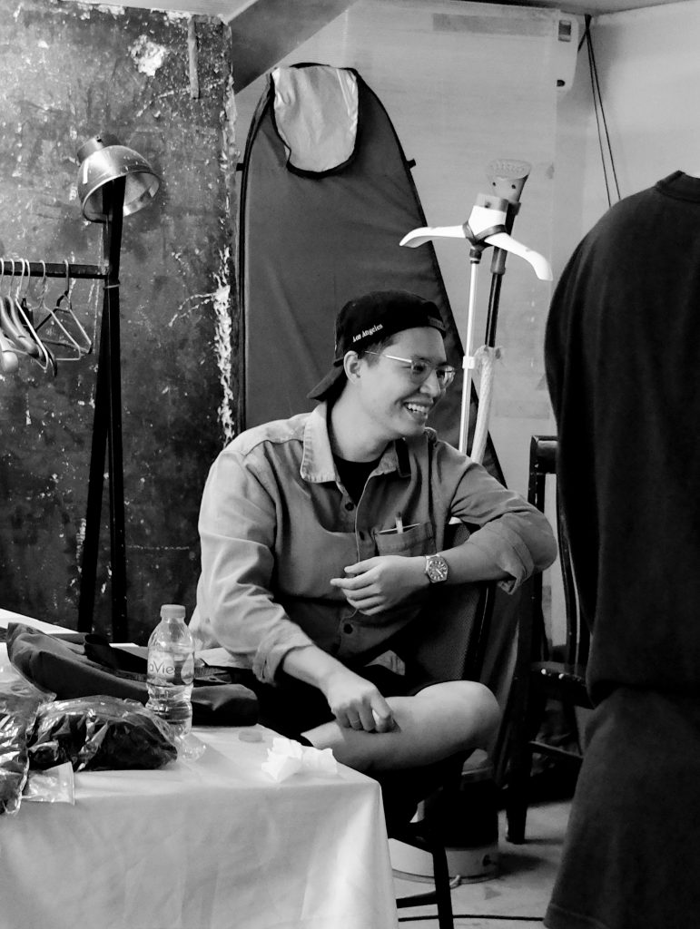 Người đàn ông đội mũ lưỡi trai đang ngồi cười rạng rỡ trong hậu trường chụp ảnh