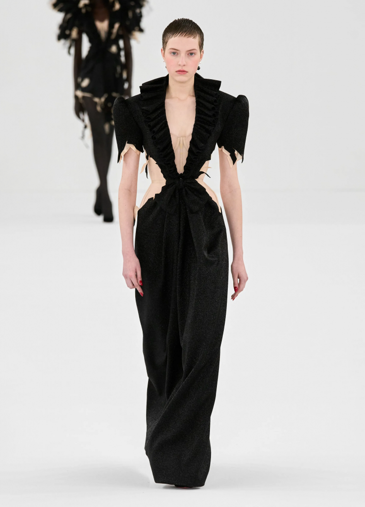 Người mẫu trình diễn váy dạ hội đen lấp lánh với cổ khoét sâu và tay áo bồng bềnh trên sàn diễn thời trang