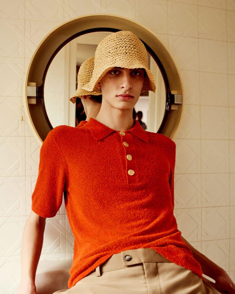 Người mẫu mặc áo polo cam đội mũ rơm nhìn vào gương tròn
