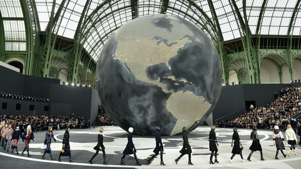 Models walk a runway circling a large globe installation inside a grand hall