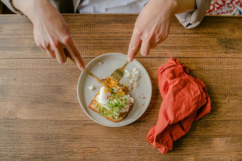 Bàn tay dùng dĩa và dao cắt bánh mì nướng kèm rau và trứng lòng đào trên đĩa