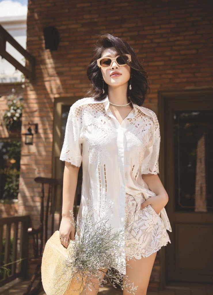 Người mẫu mặc áo sơ mi trắng ren và quần short cầm nón rơm tạo dáng ngoài trời