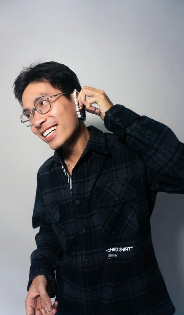 A smiling man wearing glasses and a plaid shirt holds wireless earbuds
