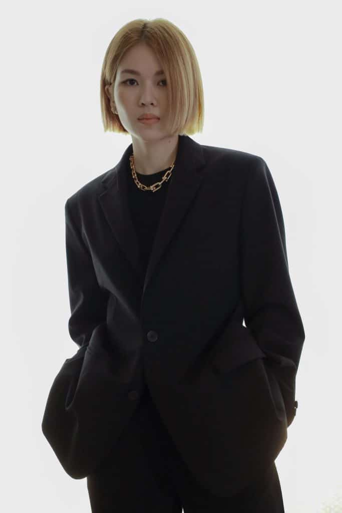 A person with blonde hair wears a large black blazer and gold chain