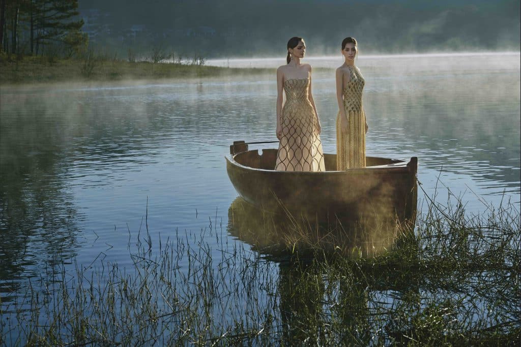Hai người mẫu diện váy dạ hội lộng lẫy trên một chiếc thuyền nhỏ giữa hồ nước sương mù
