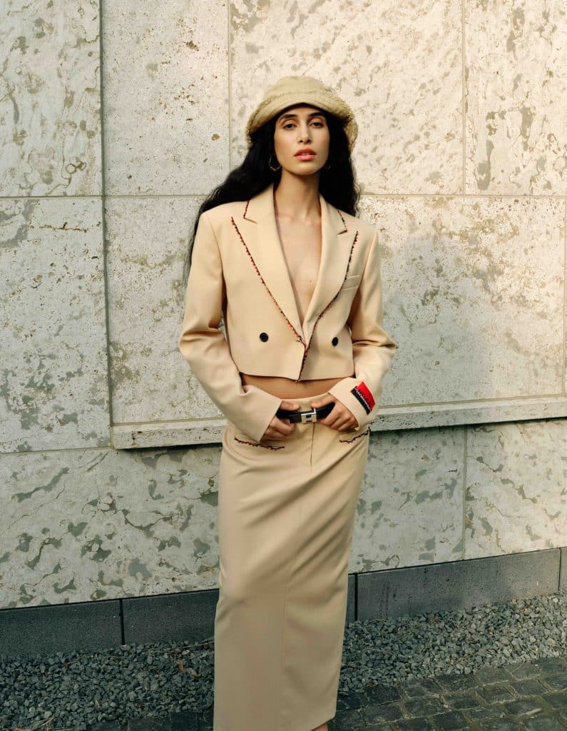 Người mẫu mặc bộ trang phục vest màu be và đội mũ đứng trước bức tường đá