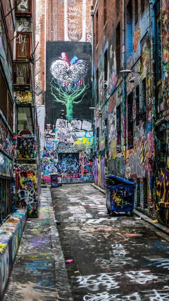 A colorful graffiti filled alleyway features a large tree mural and a blue trash bin