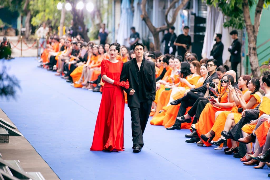 Người mẫu sải bước trên sàn diễn màu xanh lam với khán giả mặc trang phục màu cam